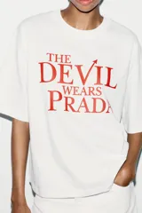 Camiseta blanca de algodón con cuello redondo, mangas cortas y hombros caídos. Estampado frontal con el título de la película "El Diablo Viste de Prada" en letras rojas.