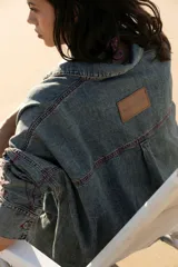 Vestido camisero de denim gris con cuello camisero, cierre frontal con botones, mangas largas con puños abotonados, bolsillos laterales y apliques bordados en el pecho y la manga. Corte holgado y largo a la rodilla.