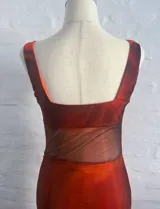 Vestido largo ajustado de microtul, color naranja con degradado a tonos rojizos y detalles en tonos morados/fucsia en la zona del torso. Presenta escote en V con efecto drapeado y tirantes anchos.