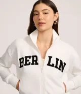 Chaqueta deportiva blanca con cierre frontal, cuello alto y puños ajustados. Presenta la inscripción "BERLIN" en letras negras y gruesas en el pecho.