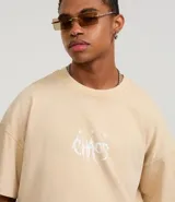 Remera de algodón color beige claro, corte boxy, con cuello redondo y mangas cortas. Presenta un estampado frontal en blanco con la palabra "Chaos" en tipografía estilizada.
