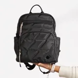 Mochila color beige con diseño acolchado en forma de rombos. Tiene un bolsillo frontal con cierre, dos bolsillos laterales abiertos y un bolsillo interno con cierre. Las correas son ajustables y tiene un asa superior para llevar en la mano.