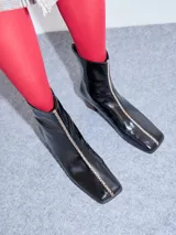 Botas de cuero charol negro con caña media y punta cuadrada. Cuentan con cierre trasero, tirador y bordado contrastante en el empeine. El taco es de madera y la suela de goma.