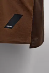 Camiseta deportiva marrón confeccionada en tejido técnico ligero y elástico con estructura perforada. Cuello redondo, manga corta y bajo asimétrico con aberturas laterales. Detalle de logo estampado.