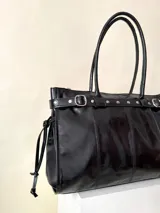 Bolso grande de cuero marrón con textura tipo cocodrilo, estructura rígida y asas para llevar al hombro. Tiene una tira con tachas y hebillas en cada extremo, también lleva lazos en los costados.