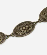 Cinturón de cadena marca Satinato confeccionado en metal con acabado envejecido. Presenta placas ovales con relieves ornamentales de flores, cierre de mosquetón y cadena de ajuste.