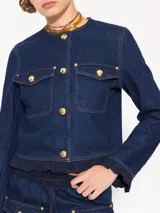 Chaqueta de denim color crudo, corte a la cintura y mangas largas con volados en puños y ruedo. Cierre frontal con broches dorados y bolsillos con broches dorados.