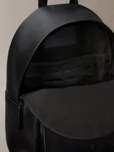 Mochila negra de diseño moderno con acabado tipo piel sintética. Presenta un asa de mano superior, correas ajustables para el hombro, compartimento principal con cremallera y un bolsillo exterior frontal con el monograma 'CK' grabado en relieve.
