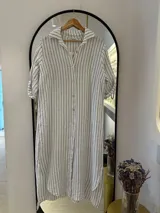 Camisola larga de lino a rayas verticales blancas y negras, con cuello camisero, cierre frontal con botones y mangas cortas con botón ajustable.