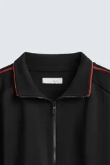 Chaqueta deportiva negra con cuello alto, cierre frontal con cremallera y ribetes rojos en hombros y mangas.