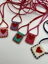 Collar con dije cuadrado bordado con un corazón rojo en el centro y bordeado con cuentas de colores. El collar es de gamuza de colores.