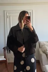 Chaqueta tipo bomber de cuero ovino marrón desgastado y gamuza color chocolate, con flecos largos en el canesú frontal y trasero. Presenta cuello solapa y cierre frontal con broches forrados.