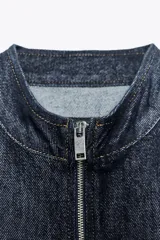 Capa corta de denim azul con cuello alto y mangas amplias. Cierre frontal con cremallera metálica plateada.