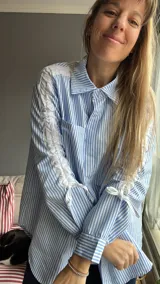 Camisa celeste a rayas verticales blancas, de corte holgado, con cuello clásico, mangas largas con ajuste de lazos y detalle de volados en los hombros.