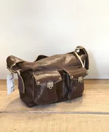 Bolso tipo hobo de cuero color marrón oscuro con acabado brillante. Tiene correa de hombro ajustable con hebilla dorada y múltiples bolsillos externos con cierre de broche metálico.