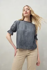 Blusa azul grisáceo de algodón con bordados frontales y terminaciones festoneadas. Tiene mangas cortas con frunces y cuello redondo con detalle de puntilla.