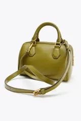 Bolso formato mini bowling color verde oliva con detalles metálicos dorados en las asas. Tiene doble asa de mano y correa bandolera extraíble. Incluye bolsillo interior con cremallera y cierre principal con cremallera.