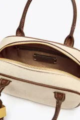 Bolso mini color crudo con detalles en marrón. Tiene asa de mano y doble asa bandolera extraíble, una en tejido tipo canvas y otra con pespunte. Incluye charm con piezas metálicas y bolsillo interior con cremallera.