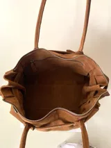 Bolso grande de cuero marrón con textura tipo cocodrilo, estructura rígida y asas para llevar al hombro. Tiene una tira con tachas y hebillas en cada extremo, también lleva lazos en los costados.