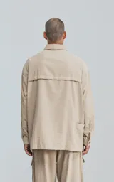 Camisa de manga larga color beige, estilo técnico con calce oversize. Presenta cuello camisero, cierre frontal de botones y múltiples bolsillos de parche con solapa en el frente, algunos con tiradores negros.