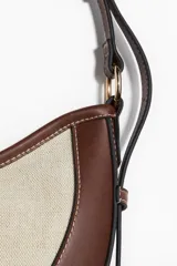 Cartera marrón con panel frontal de lona color crema, forma de media luna y correa ajustable.