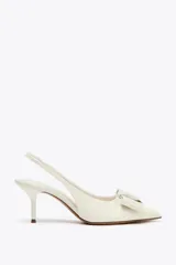 Zapato de tacón destalonado color blanco acharolado, con punta fina, lazo decorativo y tira trasera elástica.
