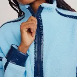 Buzo de tejido tipo sherpa color celeste con cuello alto y cierre parcial. Presenta hombros caídos, vivos en contraste azul marino, bolsillos laterales y cintura ajustable con cordón.