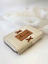 Billetera pequeña de cuero color crema, con cierre metálico y monedero interno. Presenta un diseño de cruces marrones en el frente y textura símil cocodrilo en un lateral.