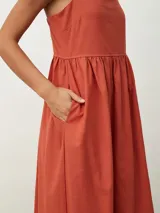 Vestido midi de color naranja, con escote recto y tirantes finos que se anudan sobre los hombros. Posee una cintura entallada con frunces que dan paso a una falda fluida.