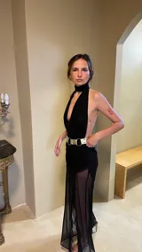 Vestido largo negro con escote profundo tipo halter y cuello alto. La falda presenta frunces y capas de microtul transparente. Se complementa con un cinturón ancho metálico dorado.