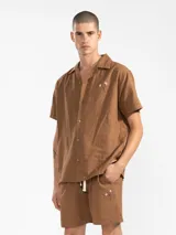 Bermuda de lino color marrón con bordado de flores blancas en la pierna izquierda. Tiene cintura elástica con cordón de ajuste, bolsillos laterales y un bolsillo trasero tipo parche.