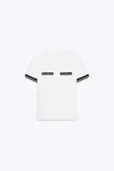 Camiseta blanca de corte recto con cuello redondo y mangas cortas. Presenta dos falsos bolsillos en el delantero con detalle de fornitura a contraste en negro y dorado.