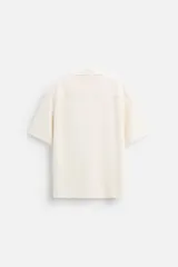 Camisa color crudo de corte regular fit, confeccionada en tejido con textura. Tiene cuello bowling, manga corta, bajo con aberturas laterales y cierre frontal con botones.