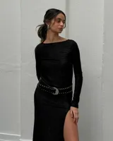Vestido largo negro de tela fina brillosa, con corte ajustado, mangas largas, cuello redondo y tajo en la pierna. Incluye cinturón de cuero negro con hebilla plateada y tachas.