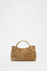 Bolso mini tipo saca de cuero serraje color marrón con efecto soft. Tiene doble asa de mano metálica dorada y asa bandolera extraíble. Cuenta con bolsillo interior con cremallera y cierre mediante fruncido y botón imantado.