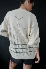 Saco de punto calado color beige claro, con cuello redondo y cierre frontal de botones azules. Presenta detalles decorativos en crochet verde y negro en el frente y puños.