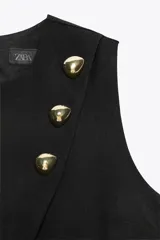 Chaleco negro con cuello redondo y sisa. Detalle de falsos botones dorados delanteros. Bajo con abertura frontal. Cierre en espalda con abertura y botón.
