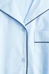 Camisa oversized de algodón color celeste claro con ribetes en contraste negro. Presenta cuello abierto, cierre frontal de botones, un bolsillo superior de parche y mangas largas con puños anchos.