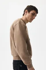 Buzo de tejido pesado de algodón con efecto lavado, color marrón topo. Presenta un diseño estampado en el frente con el texto "RIVERSIDE SOCIAL CLUB" y un escudo con un remo. Tiene cuello redondo, hombros caídos y puños y bajo acanalados.