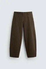 Pantalón marrón con finas rayas verticales negras, de corte amplio tipo balloon fit, con bolsillos laterales y traseros ocultos. Cierre frontal con cremallera y botonadura cruzada.