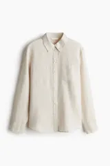 Camisa color crudo de lino liviano, con cuello clásico, botones al frente, canesú de doble capa en la espalda, hombros apenas caídos, mangas largas con puños abotonados, bolsillo superior de parche y bajo levemente redondeado.