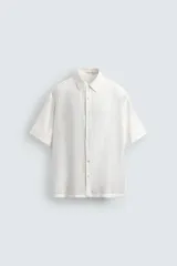 Camisa blanca de manga corta con cuello solapa y textura jacquard irregular. Corte relajado (relaxed fit) con aberturas laterales y cierre frontal de botonadura.