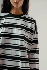 Camiseta de manga larga, corte holgado, con cuello redondo y estampado de rayas horizontales anchas en blanco y negro. Presenta un pequeño bordado rojo en el pecho.