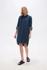 Vestido camisero de jean azul oscuro, corte holgado, largo a la rodilla, con cuello camisero, mangas largas abullonadas con puños abotonados y cierre frontal con botones.
