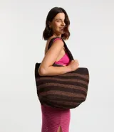 Bolso de playa tipo tote, confeccionado en paja color marrón con rayas horizontales negras. Tiene doble asa de hombro y cierre por broche a presión. Incluye un pequeño bolsillo interior.