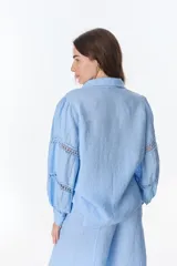 Camisa celeste de lino con cuello clásico, abotonada al frente y mangas largas con detalle de puntilla en los hombros.