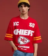 Remera roja de algodón con estampado del logo de los Kansas City Chiefs, el nombre del equipo y el número 60 en blanco. Tiene mangas cortas con rayas amarillas y rojas.