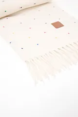 Pashmina color beige con pequeños puntos bordados en varios colores (rojo, azul, amarillo, verde, violeta). Tiene flecos en los extremos y un parche de cuero sintético marrón con un logo grabado.