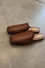 Mules de cuero color marrón con capellada tejida y suela plana.