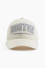 Gorra de sarga color crema con bordado azul marino de la palabra "BOSTON" y debajo "MASSACHUSETTS". Tiene botón en la parte superior, cinta antitranspirante y correa ajustable en la parte trasera.